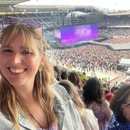 Billede af kvinde der smiler til en koncert med mange mennesker i baggrunden og storskærm med Taylor Swift på