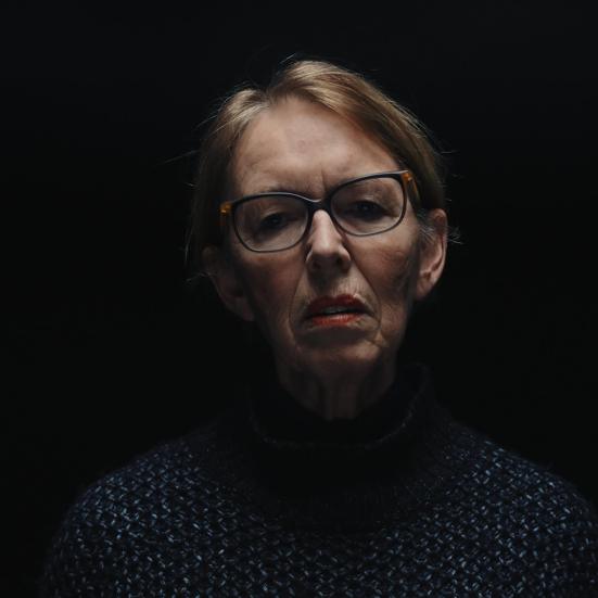 En kvinde foran en sort baggrund, hun har briller og hendes ansigt er belyst oppefra