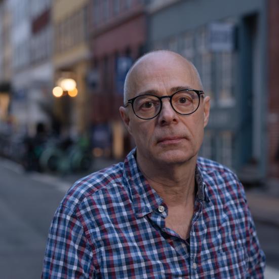 En mand med briller og skjorte på, der står ude på gaden, der bygninger i baggrunden.