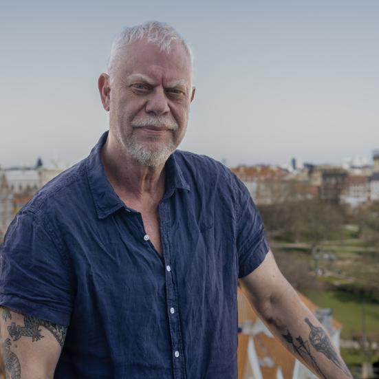 En mand med blå skjorte på, han ser ind i kameraet og der en by og noget natur i baggrunden 