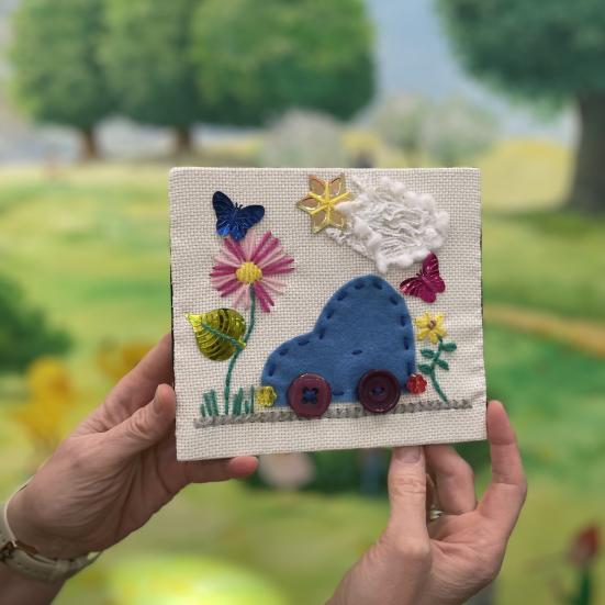 Hænder der holder et broderi billede med en bil, blomst, sol og sky på
