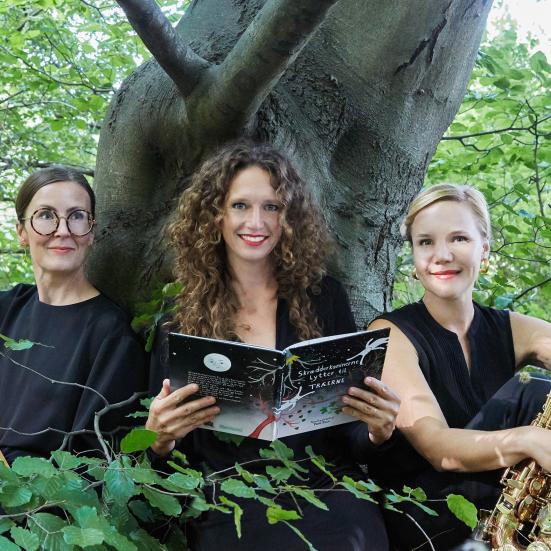 Tre kvinder der sidder foran et træ i en skov med en bog og et instrument