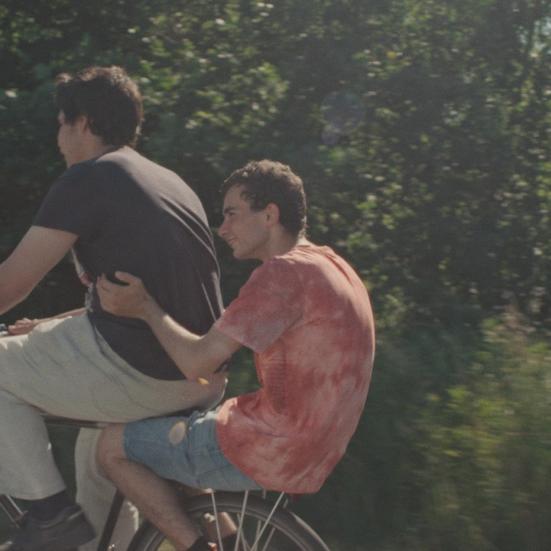 To mænd der cykler på en cykel den ene sidder bagpå og der skov i bagrunden