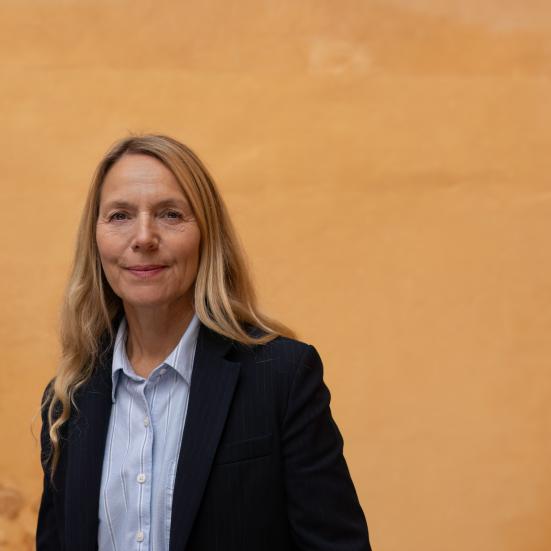 En kvinde der står og ser ind i kammeret, med sort blaser og lys skjorte foran en orange baggrund