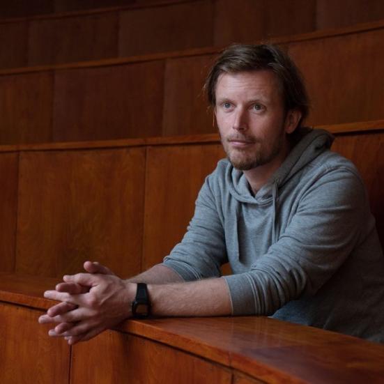 Mand der sidder i et lokale med træmøbler, med folede hænder og ser frem for sig.