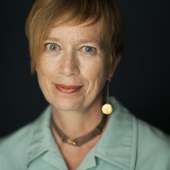 Kvinde med kort hår foran sort baggrund, med halskæde og blå skjorte, der ser lige ind i kameraet