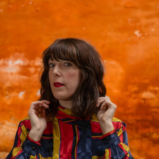 Kvinde foran en orange murstensmur i en farvestålende jakke
