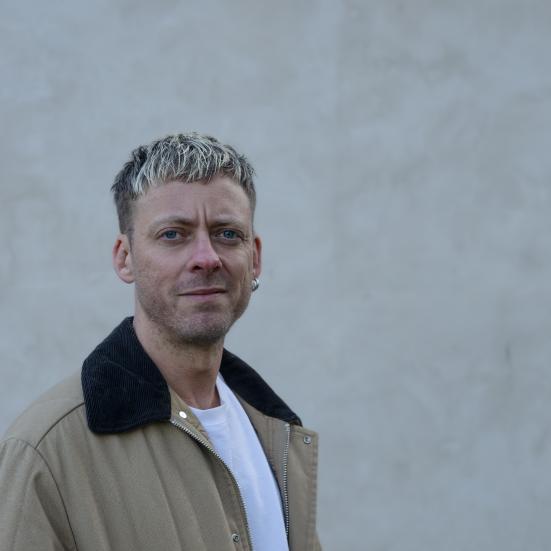 Mand der står foran en grå murstensvæg og ser lige ind i kammaret med en jakke på