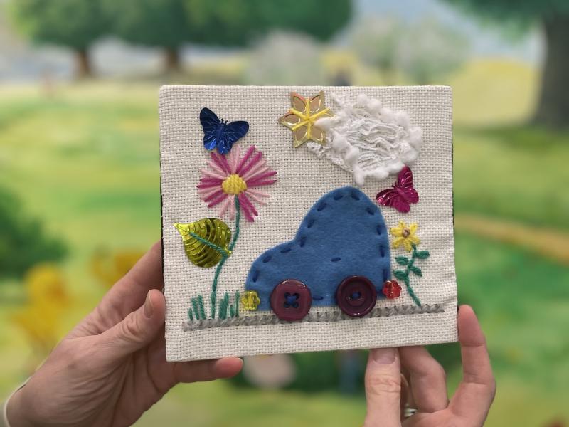 Hænder der holder et broderi billede med en bil, blomst, sol og sky på