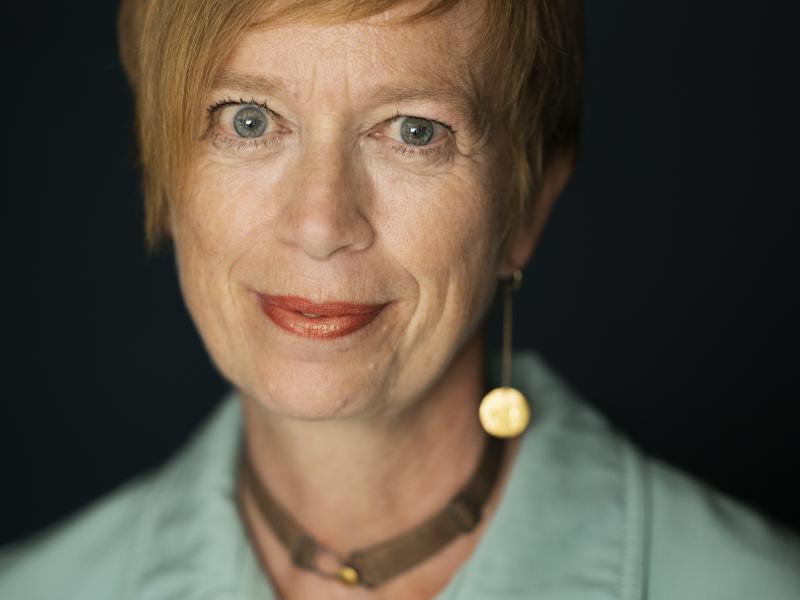 Kvinde med kort hår foran sort baggrund, med halskæde og blå skjorte, der ser lige ind i kameraet