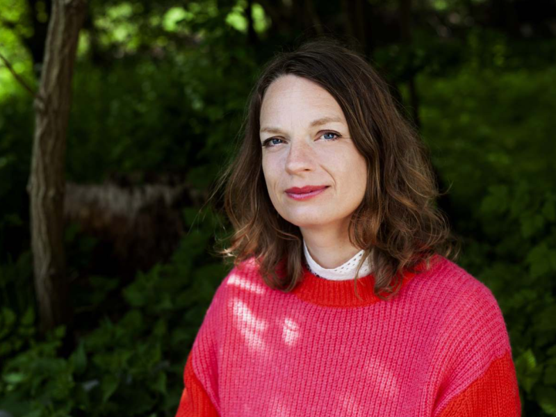 Et billede af Charlotte Weitze i en rød sweater med træer i baggrunden.