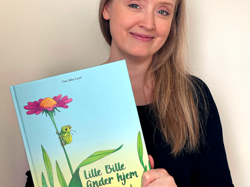 Kvinde med langt lyst hår holder en blå bog med en blomst og græs på