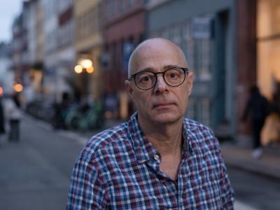 En mand med briller og skjorte på, der står ude på gaden, der bygninger i baggrunden.