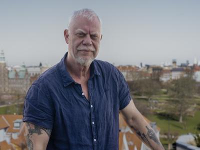 En mand med blå skjorte på, han ser ind i kameraet og der en by og noget natur i baggrunden 