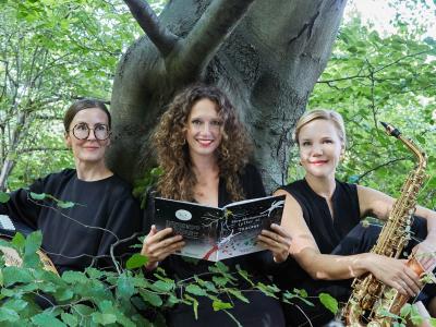 Tre kvinder der sidder foran et træ i en skov med en bog og et instrument