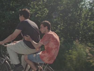 To mænd der cykler på en cykel den ene sidder bagpå og der skov i bagrunden