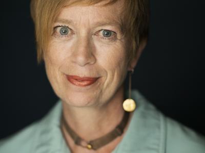 Kvinde med kort hår foran sort baggrund, med halskæde og blå skjorte, der ser lige ind i kameraet