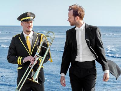 To mand der står ved havet iført sort tøj og hvide skjorter den ene har et blæser instrument i hænderne