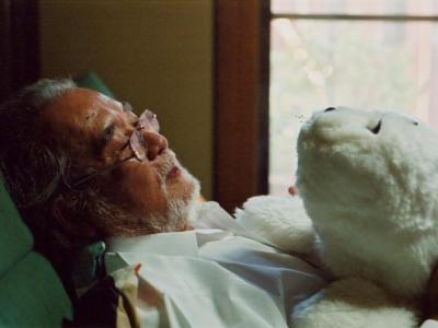 Ældre mand der ligger ned i en seng med en mekanisk bamse på brystet