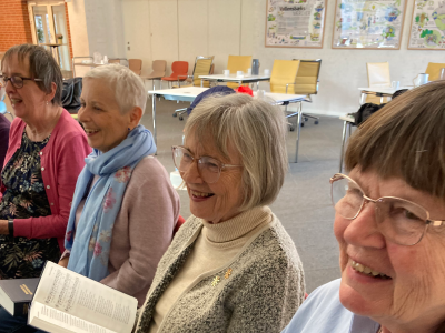 4 smilende ældre damer sidder med åbne sangbøger i et veloplyst lokale