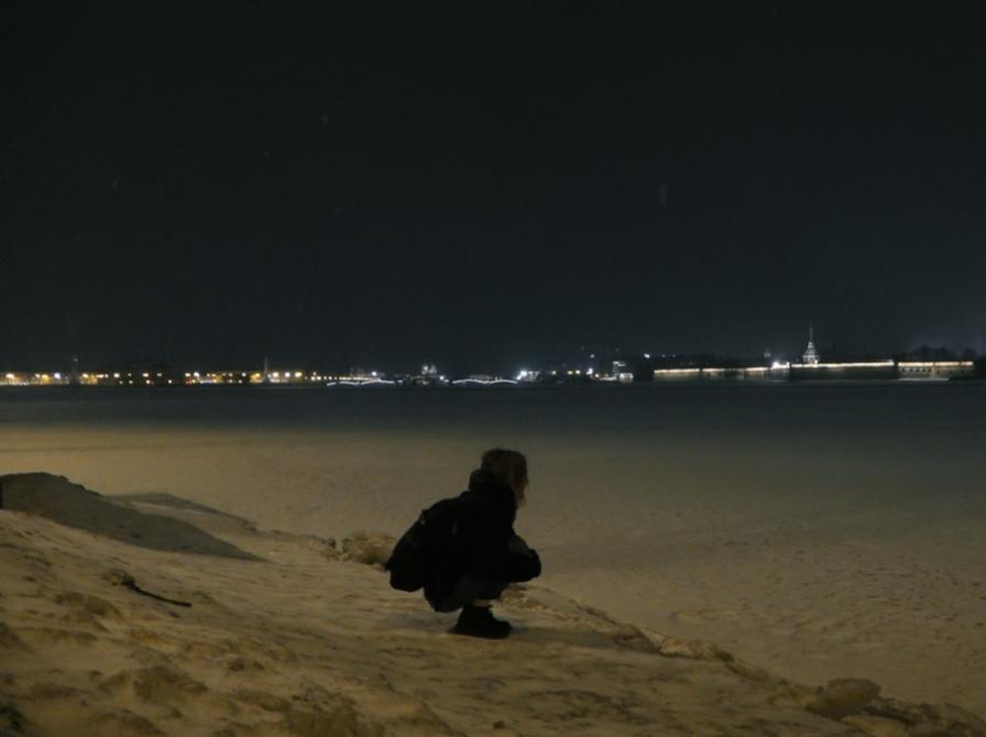 En skikkelse sidder på hug, i sandet om aften med en by i baggrunden