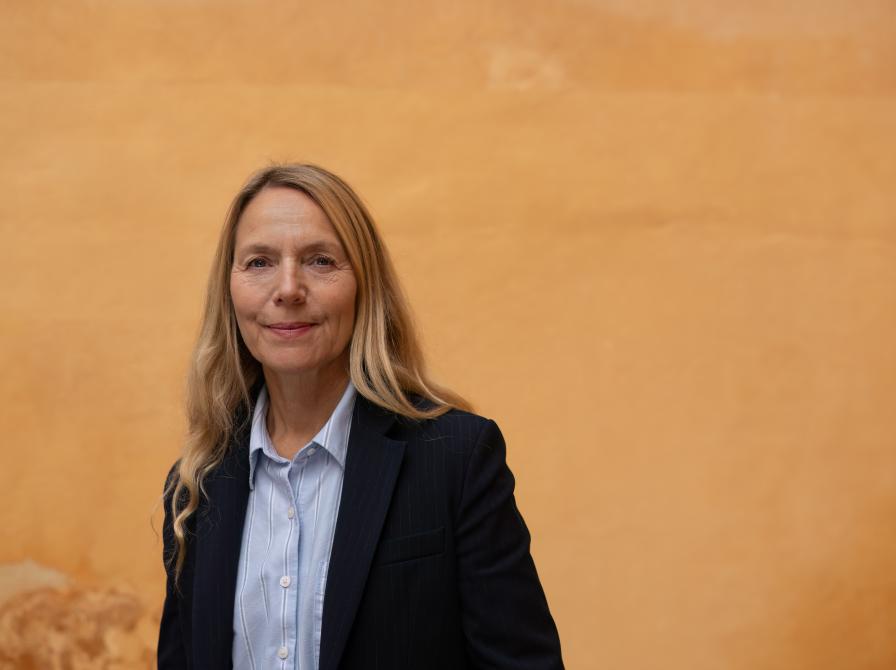 En kvinde der står og ser ind i kammeret, med sort blaser og lys skjorte foran en orange baggrund