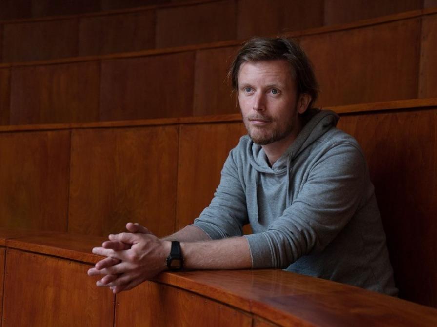 Mand der sidder i et lokale med træmøbler, med folede hænder og ser frem for sig.