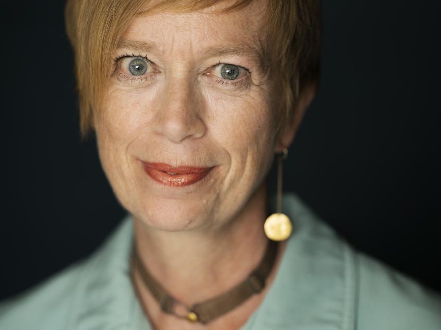 Kvinde med kort hår foran sort baggrund, med halskæde og blå skjorte, der ser lige ind i kameraet