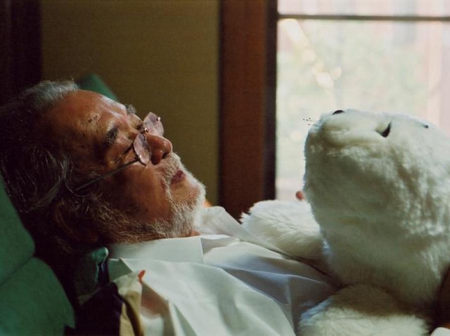 Ældre mand der ligger ned i en seng med en mekanisk bamse på brystet