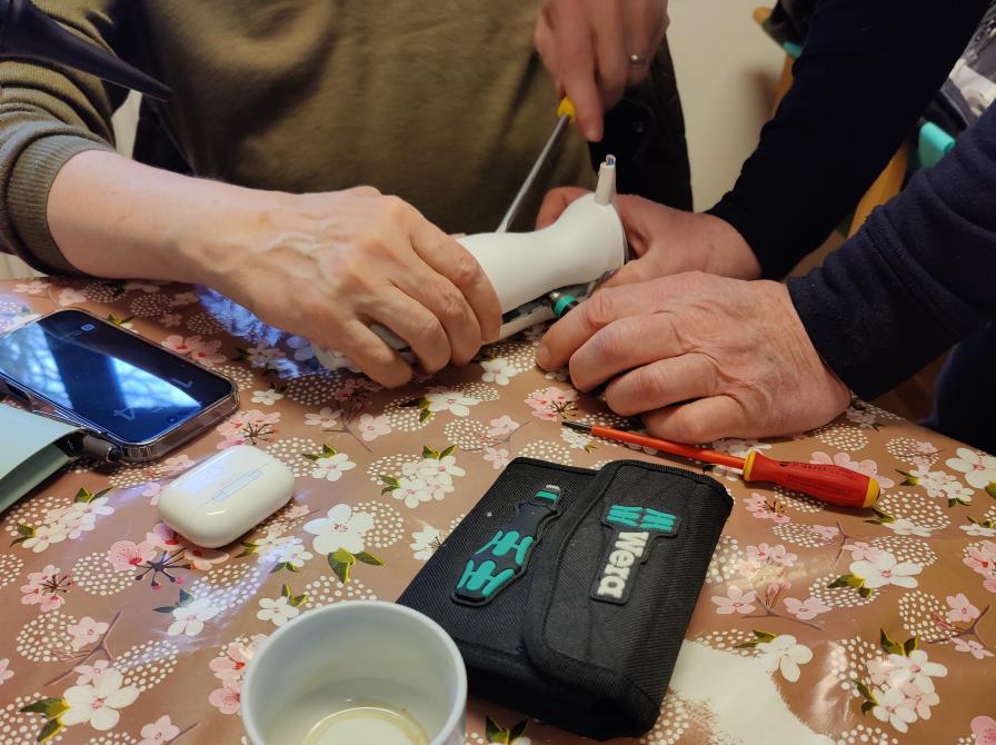 billede af et bord hvorpå der ligger flere værktøjer til arbejde med elektronik, to par hænder er i gang med at arbejde på en stavblender